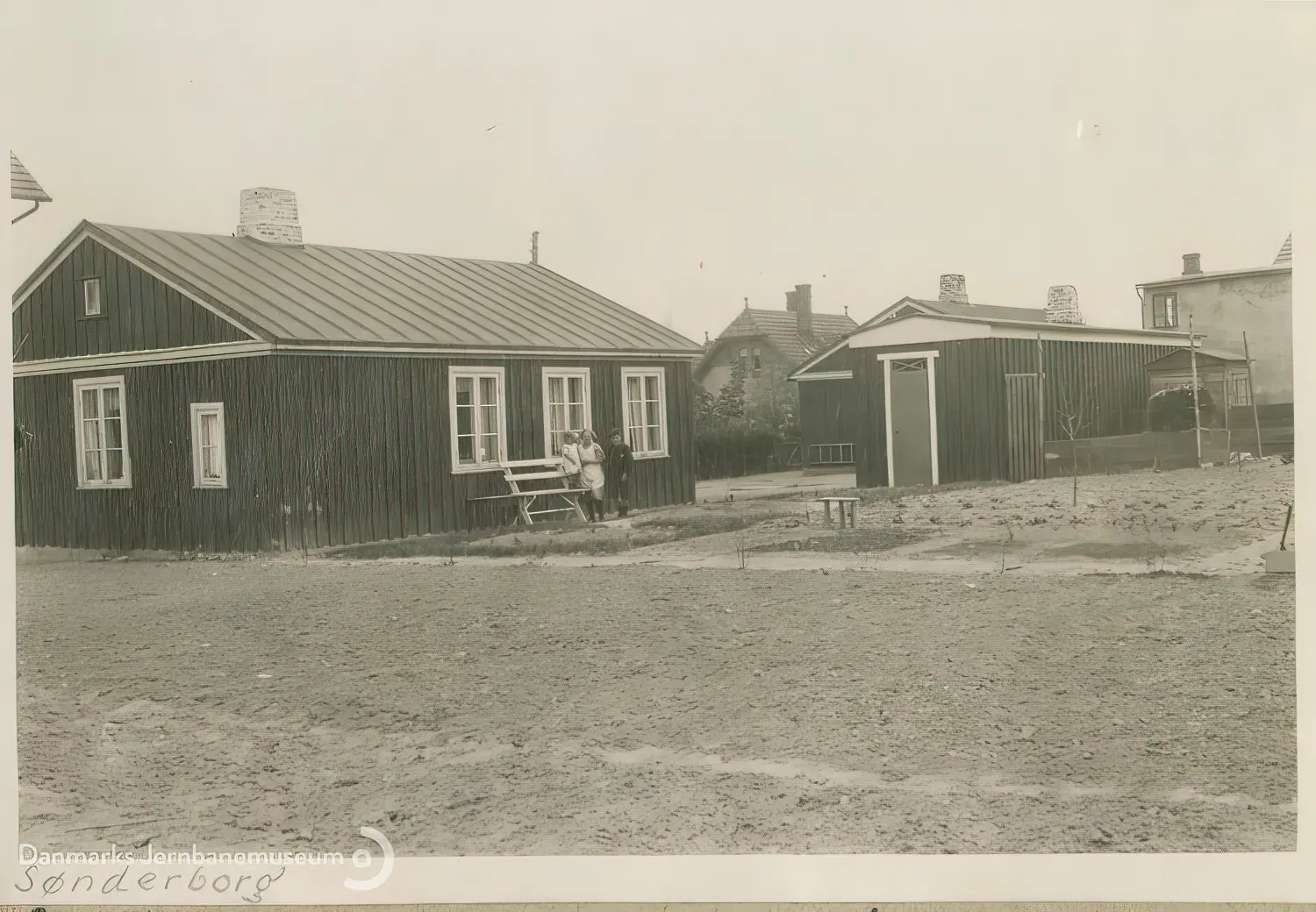 Billede af midlertidige boliger for jernbanefolk til Sønderborg Hovedbanegård efter Genforeningen. Havesiden af svellehusene på Skrænten 25 og 26. I midten en fælles retiradebygning.
