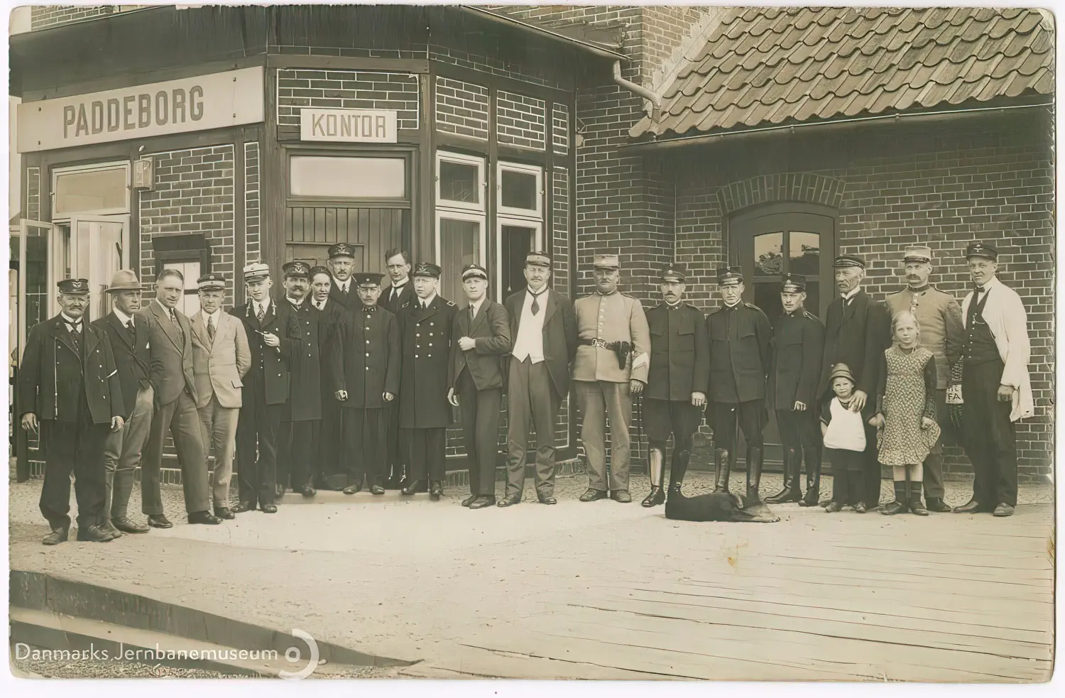 Billede af civile, toldere, politi, gendarmer og en enkelt tysk tjenestemand på perronen under det nye danske stationsnavneskilt efter den danske overtagelse af Padborg Station natten mellem den 16. og 17. jun. 1920.